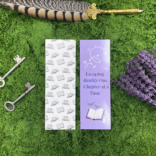 Two sides of a bookmark shown side by side, one purple with a glowing open book, bats, and stars reading "Escaping Reality One Chapter at a Time," the other white with a repeating pattern of small open books and stars, resting on artificial grass next to two vintage keys, dried lavender, and a gold quill pen.