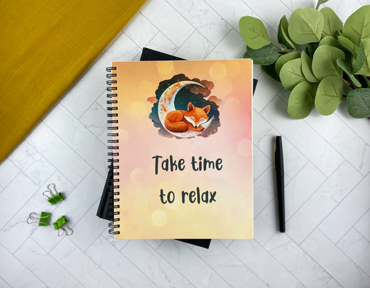 Notebook cover illustrated with a sleeping fox curled into a crescent moon against a pink-orange sunset gradient, with the words "Take time to relax," resting on a black book with green eucalyptus nearby.