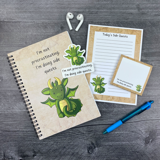Flat lay of the “I’m Not Procrastinating, I’m Doing Side Quests” stationery set on a dark grey wood surface. Includes a lined notebook, 5x7 notepad, sticky notes, and a vinyl sticker with matching text. White wireless earbuds and a blue pen are styled alongside the set to create a casual, modern desk aesthetic.
