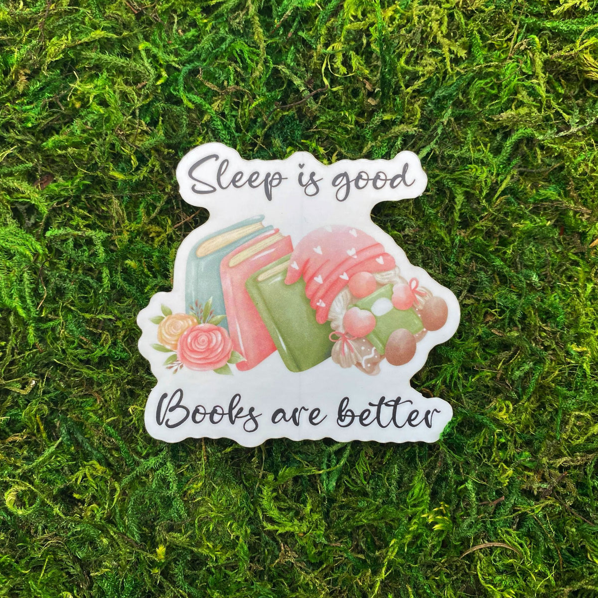 Die-cut sticker with stacked books, a sleeping gnome cradling a book, and the text 'Sleep is good, books are better,' laid on faux moss