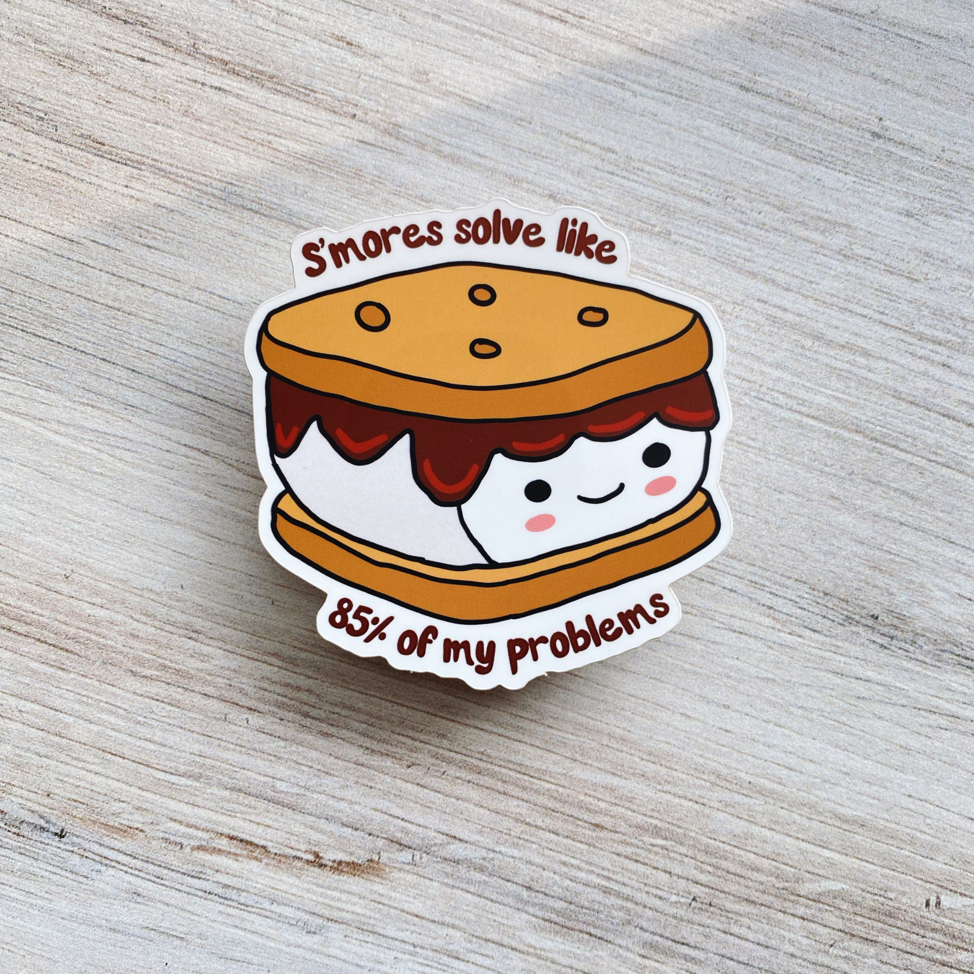 Die-cut vinyl sticker showing a smiling s'more with graham crackers and marshmallow, with the text "S'mores solve like 85% of my problems," on a light wood background