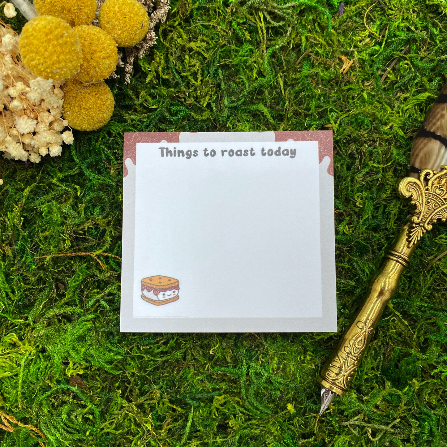Square sticky note pad with a dripping chocolate marshmallow-style white border, a small s'more illustration, and the words "Things to roast today," resting on moss next to dried yellow and white flowers and a gold quill pen.