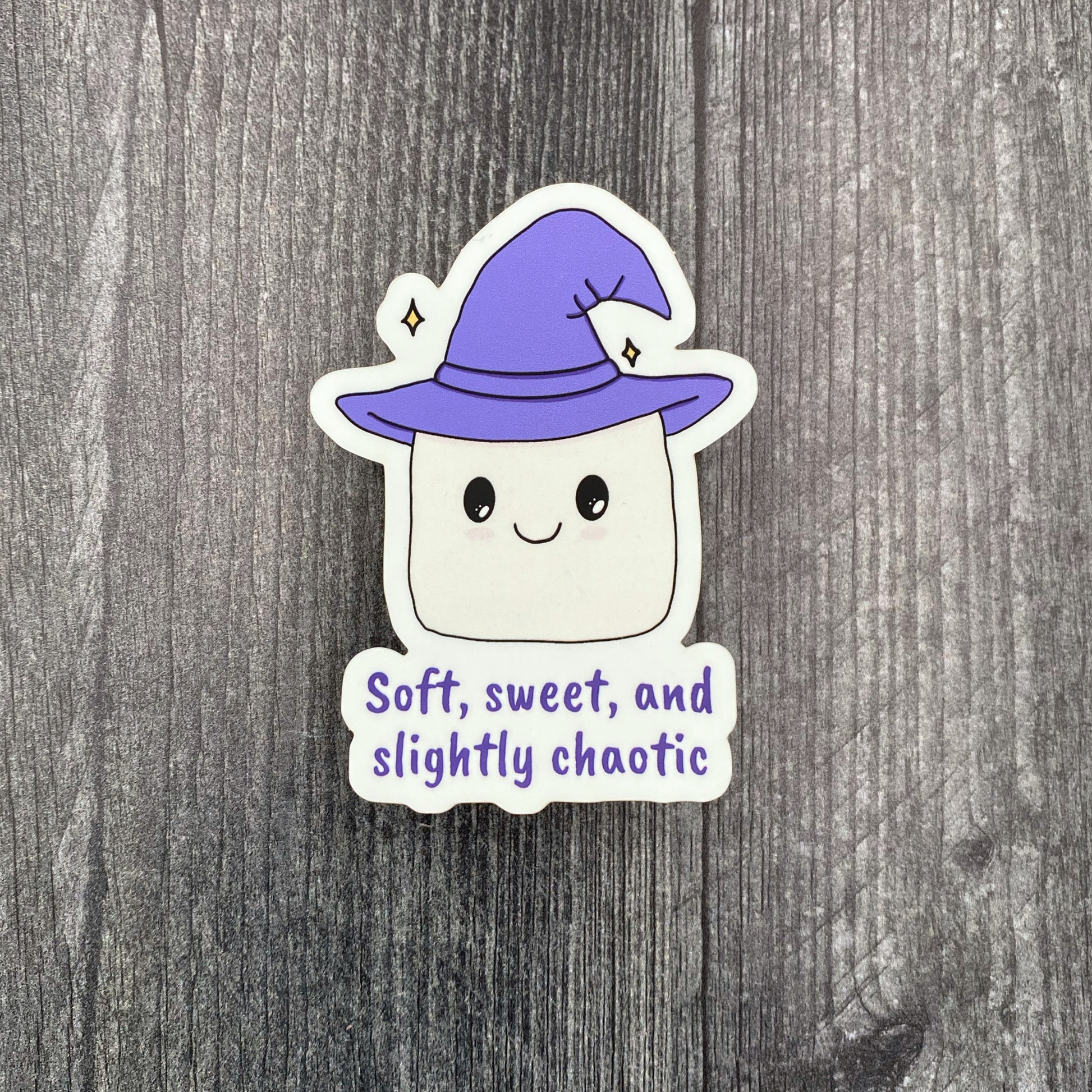 Die-cut vinyl sticker showing a smiling marshmallow wearing a purple witch hat, with the text "Soft, sweet, and slightly chaotic," on a gray wood background