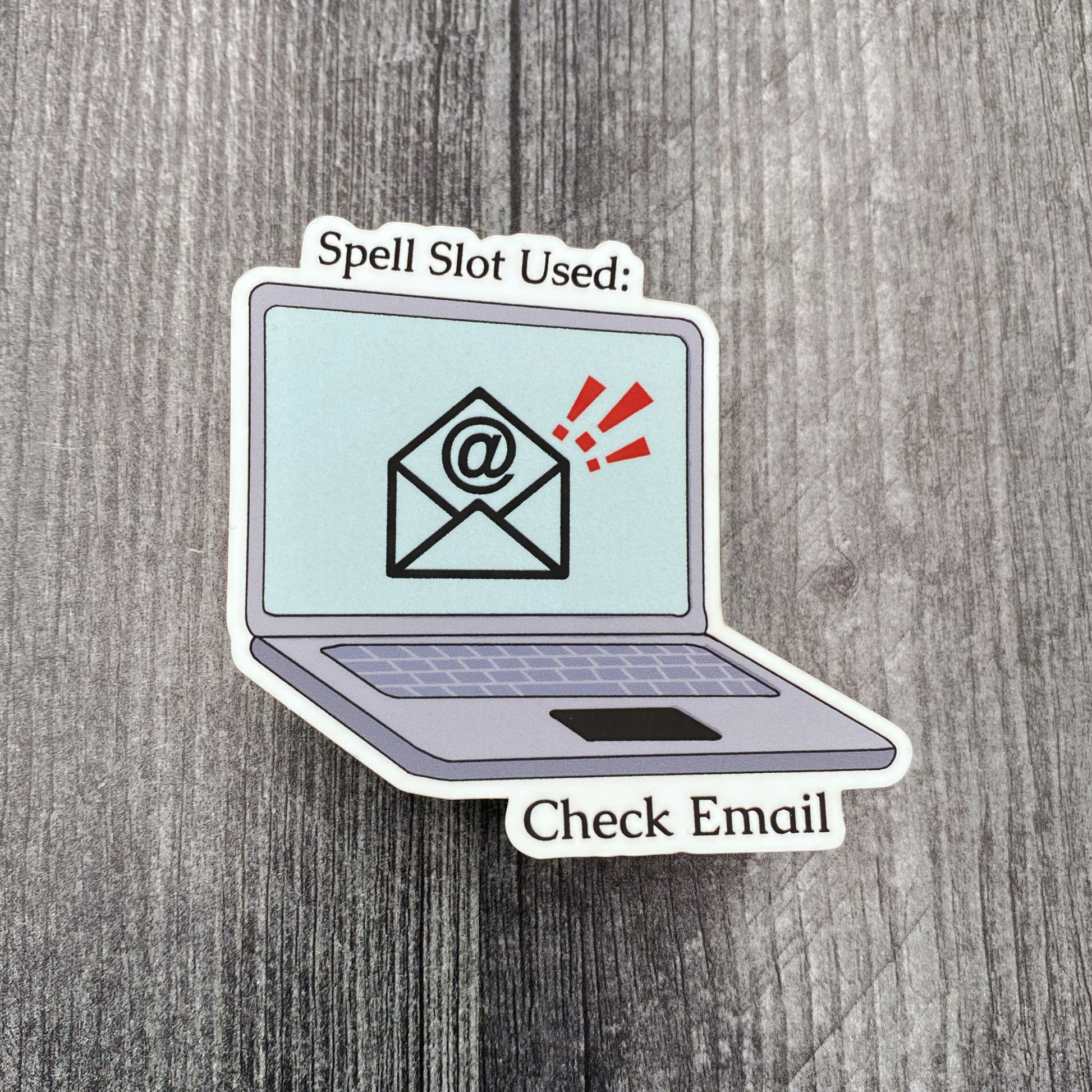 Die-cut vinyl sticker showing a laptop with an email notification, with the text "Spell slot used: check email," on a gray wood background