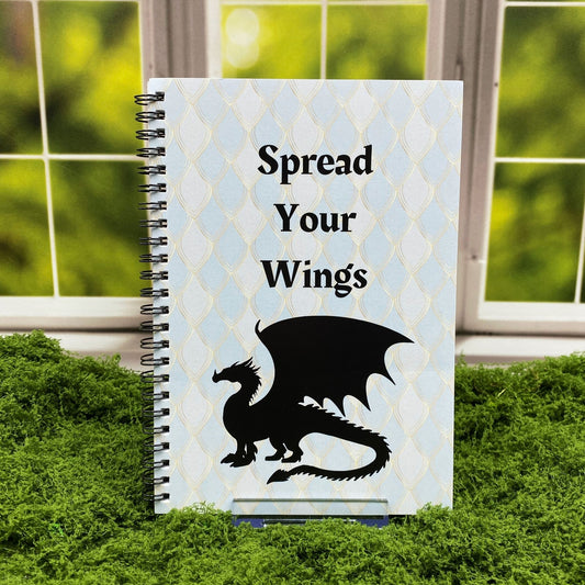 Spiral-bound notebook with a pale blue dragon-scale patterned cover reading 'Spread Your Wings' above a black dragon silhouette, displayed on a moss mat with a window backdrop.