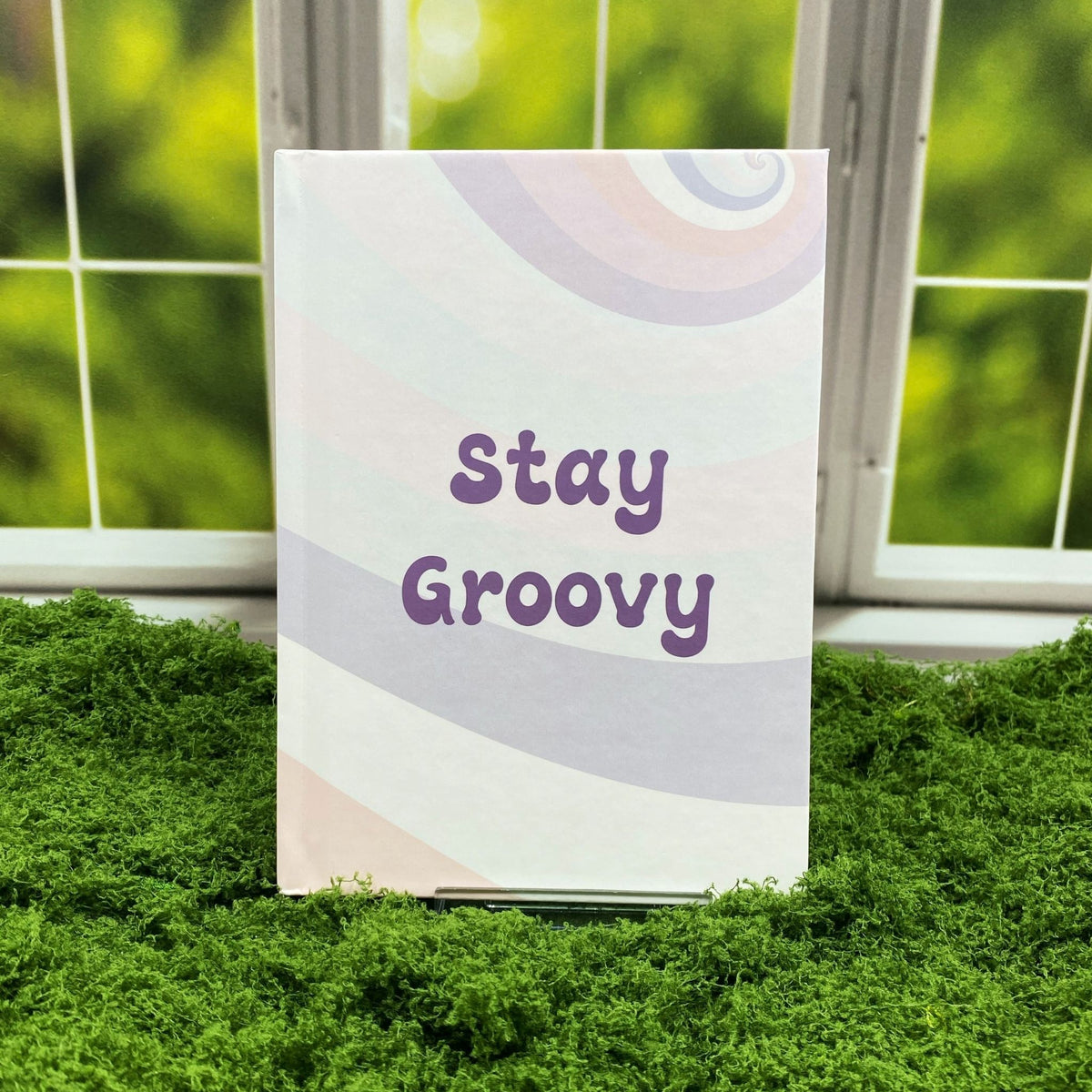 Hardcover journal with a pastel retro rainbow swirl pattern, reading 'Stay Groovy' in purple lettering, displayed on a moss mat with a window backdrop.
