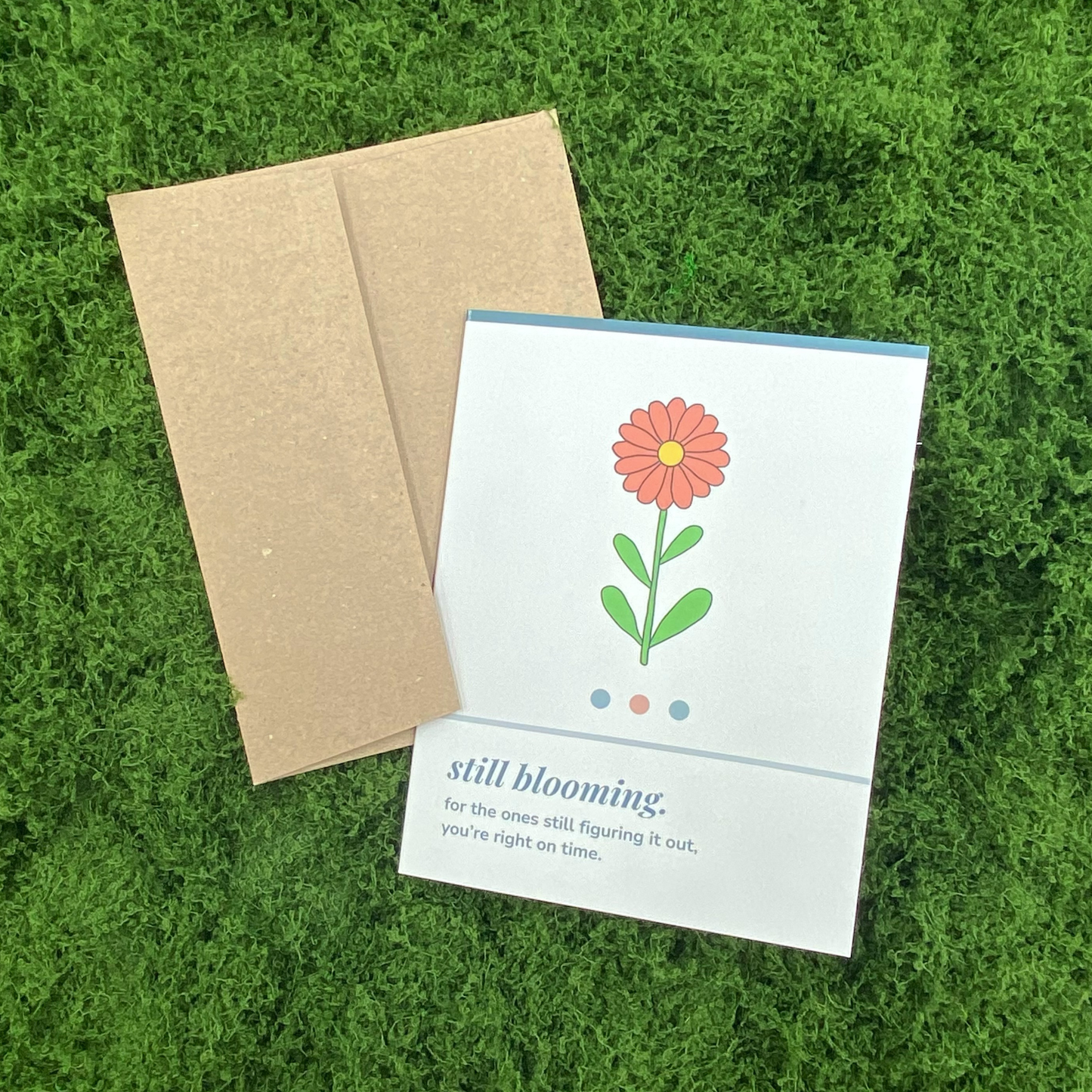 Greeting card that reads "still blooming. for the ones still figuring it out, you're right on time." with an illustration of a single orange flower, resting against its Kraft paper envelope on artificial grass.