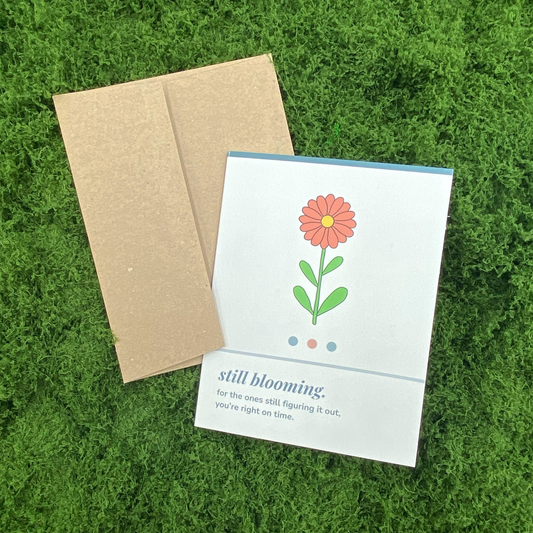 Greeting card that reads "still blooming. for the ones still figuring it out, you're right on time." with an illustration of a single orange flower, resting against its Kraft paper envelope on artificial grass.
