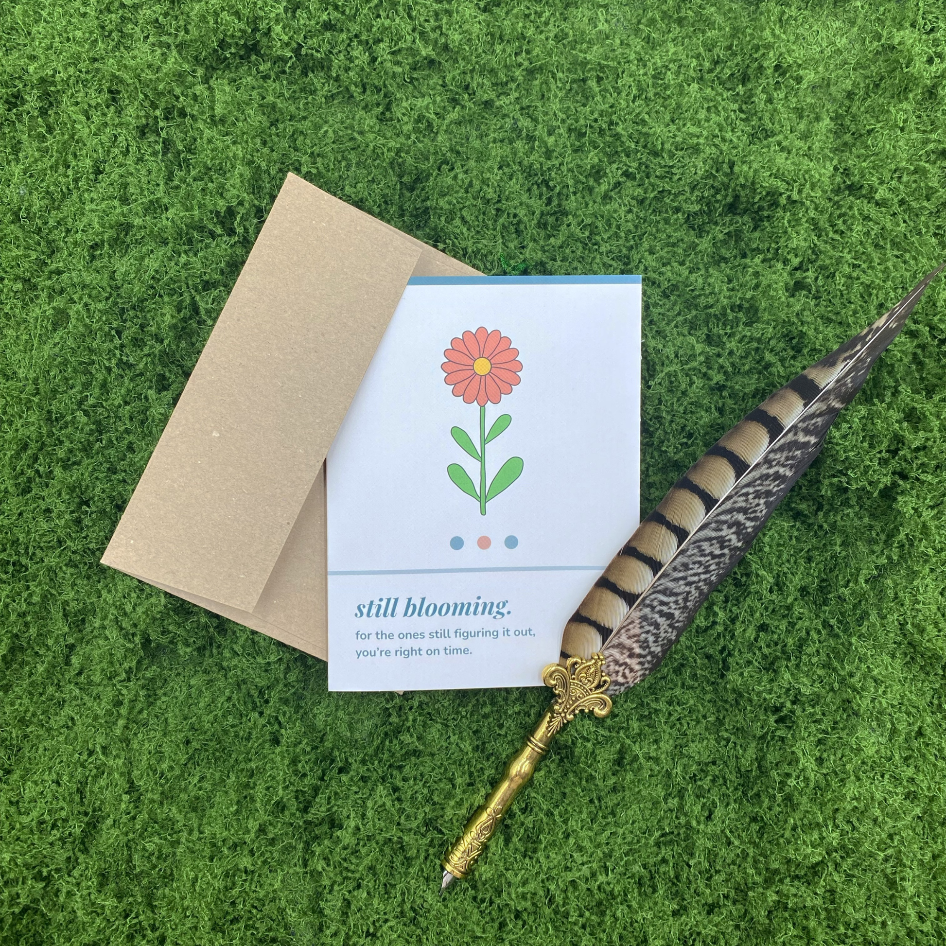 A2 matte encouragement greeting card featuring whimsical wildflower illustration with the text "still blooming — for the ones still figuring it out, you're right on time," with a kraft envelope, on a green moss background.