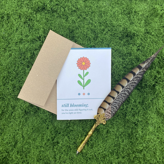 A2 matte encouragement greeting card featuring whimsical wildflower illustration with the text "still blooming — for the ones still figuring it out, you're right on time," with a kraft envelope, on a green moss background.