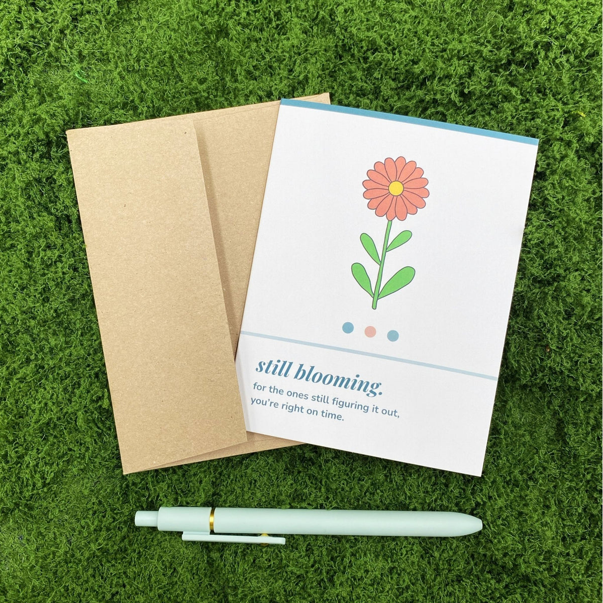 "Still Blooming" greeting card lying on a kraft brown envelope on grass, with a mint green pen beside it for scale.