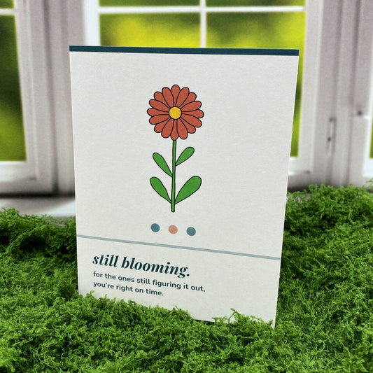 "Still Blooming" greeting card standing upright on a moss mat in front of a window backdrop, featuring an illustrated orange daisy with the printed message "for the ones still figuring it out, you're right on time."