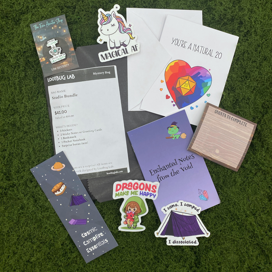 A Lootbug Lab studio bundle mystery bag with a reveal card surrounded by sample items including stickers, a bookmark, a pocket notebook, sticky note pads, and a greeting card on a green moss surface