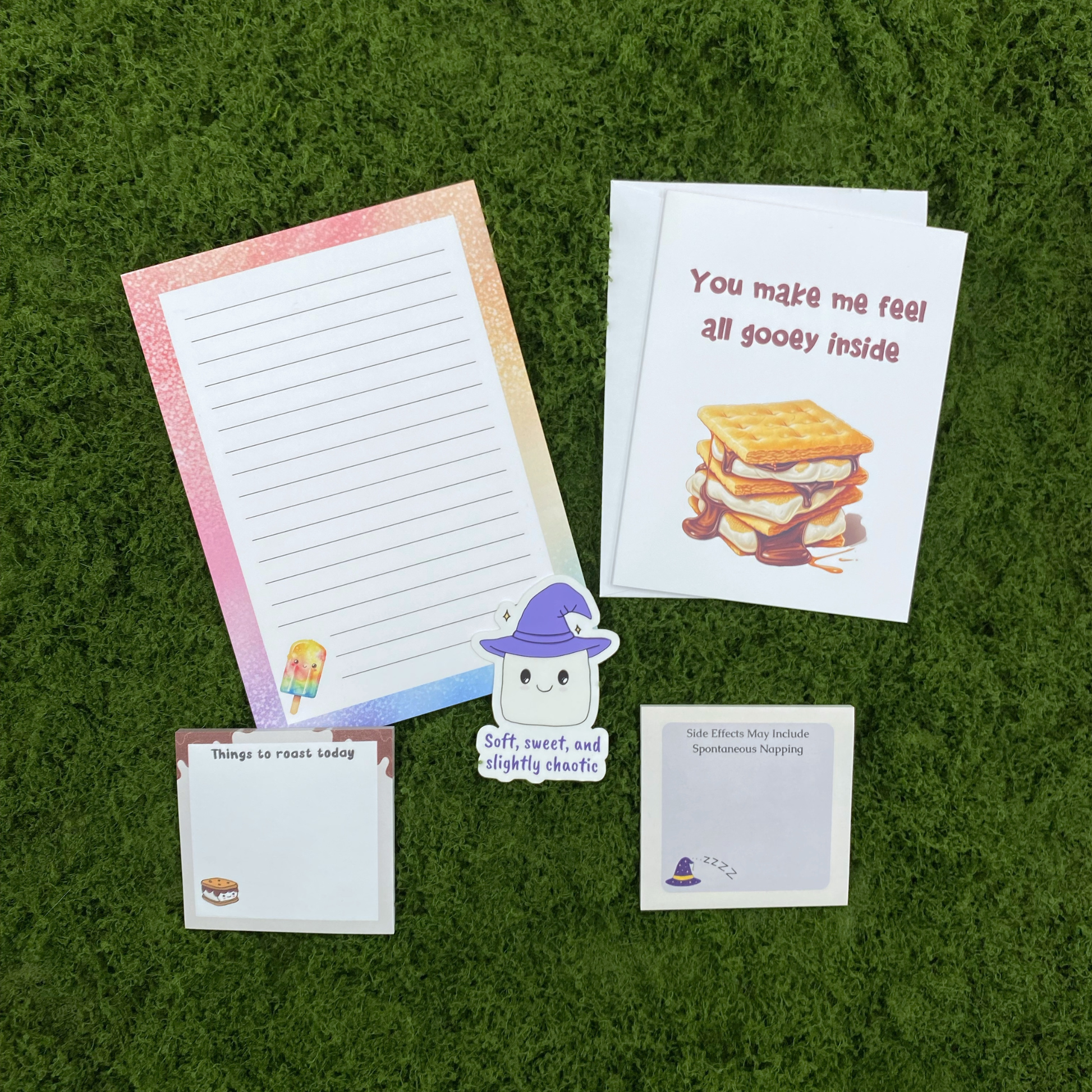 A best friend gift set including a rainbow glitter popsicle notepad, a things to roast today sticky note pad, a side effects may include spontaneous napping sticky note pad, a soft sweet and slightly chaotic marshmallow sticker, and a you make me feel all gooey inside greeting card with envelope, arranged on a green moss surface
