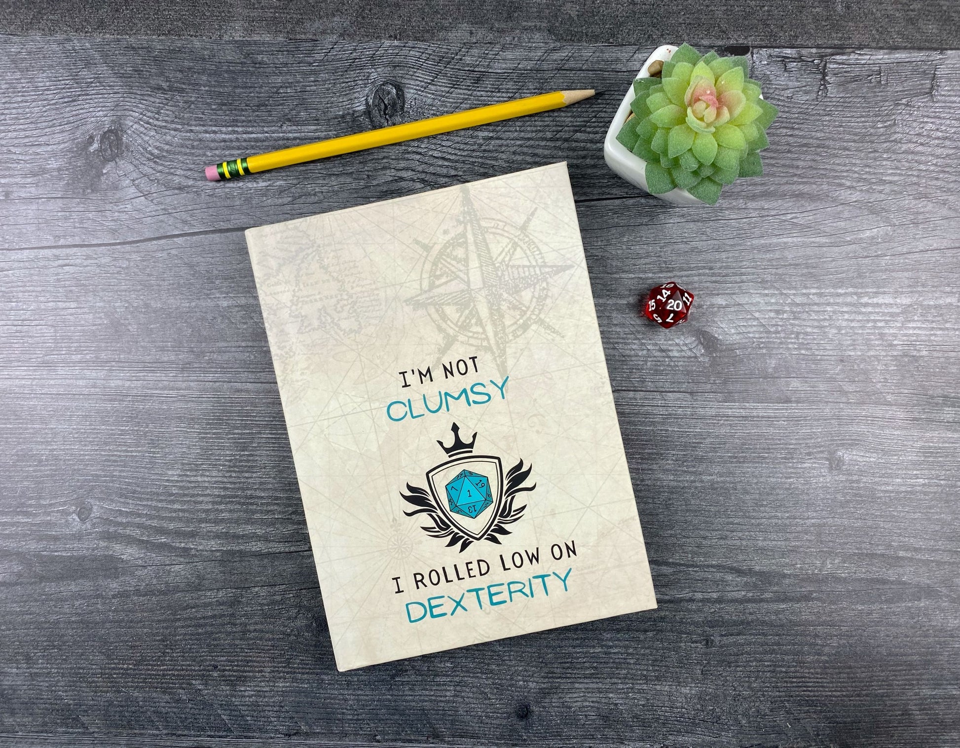 Hardcover journal with a tan parchment-and-compass design, a crowned shield with a teal d20 inside, and the words "I'm not clumsy, I rolled low on dexterity," resting on a grey wood surface next to a red d20 die, small succulent, and yellow pencil.