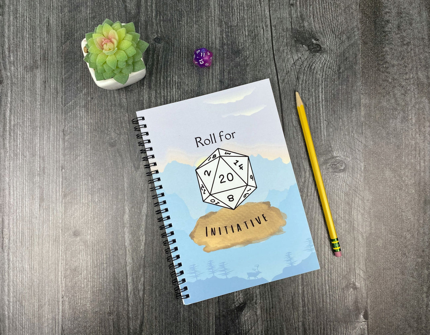 Spiral notebook cover illustrated with a white d20 die reading "Roll for Initiative" over a blue mountain and forest scene, resting on a grey wood surface next to a purple twenty-sided die, small succulent, and yellow pencil.