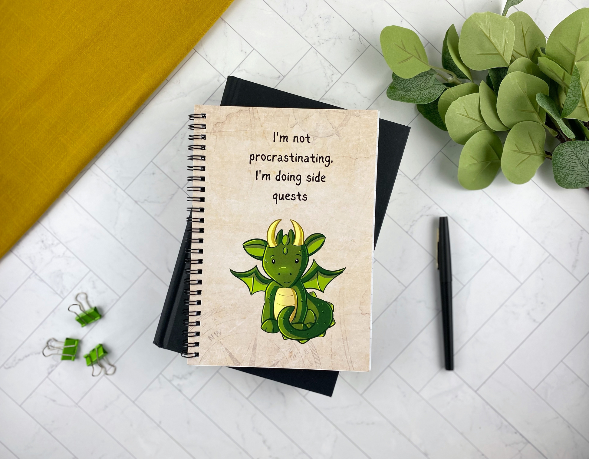 Spiral notebook cover with a tan map-textured background, illustrated with a cartoon green baby dragon and the text "I'm not procrastinating, I'm doing side quests," resting on a black notebook with green binder clips, a black pen, and eucalyptus greenery nearby.
