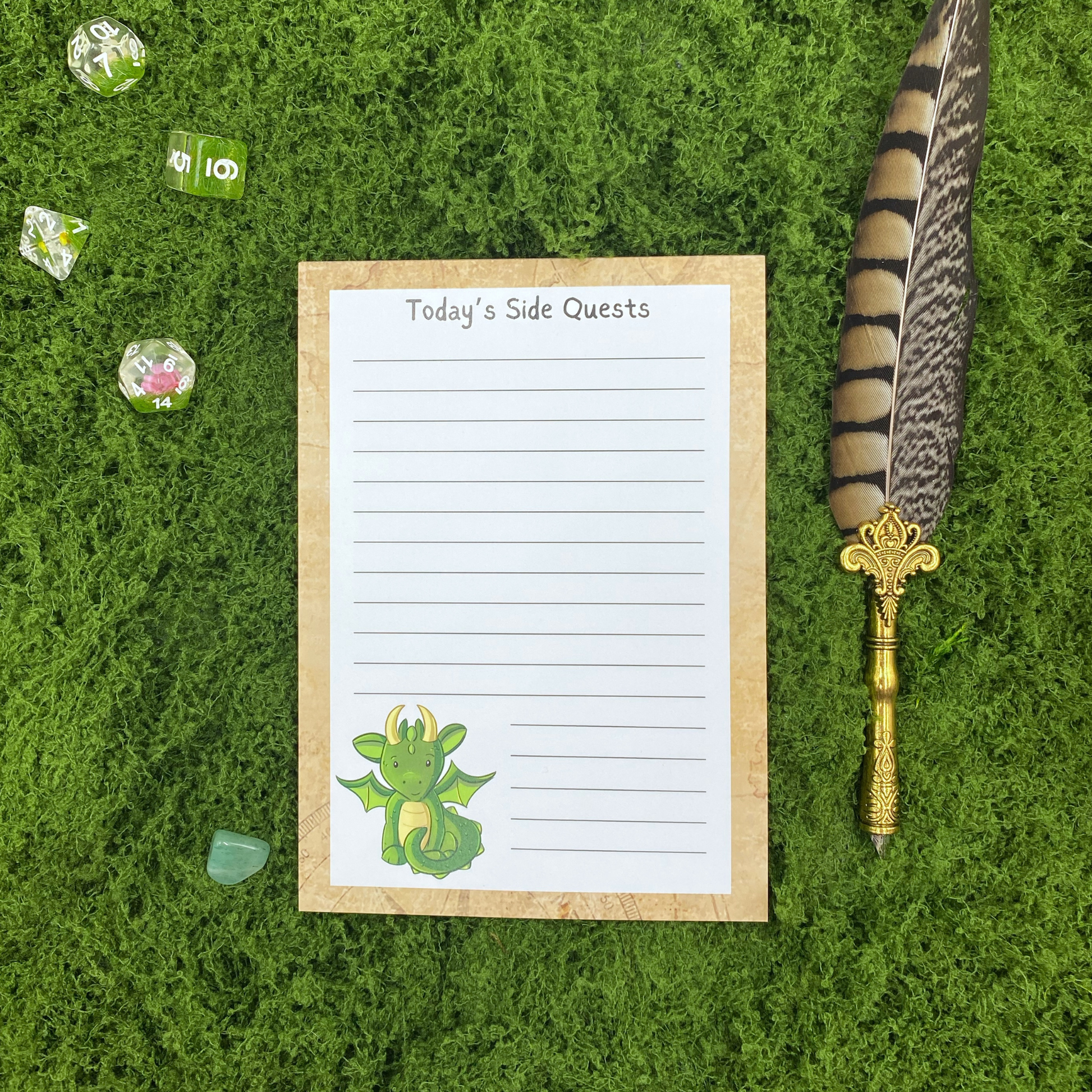 Lined notepad on a parchment-textured border headed "Today's Side Quests," with a small green dragon illustration in the corner, resting on artificial grass next to dice, a crystal, and a gold quill pen.
