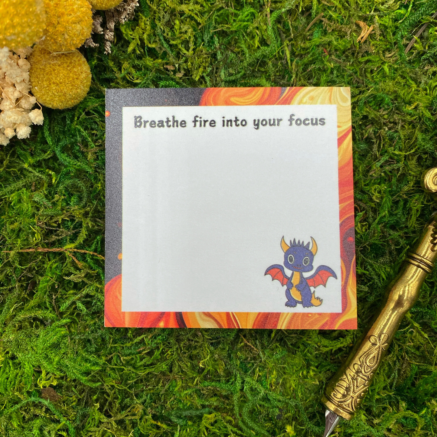 Square sticky note pad with an orange and red flame-swirl border, a small black dragon illustration, and the words "Breathe fire into your focus," resting on moss next to dried yellow and white flowers and a gold quill pen.