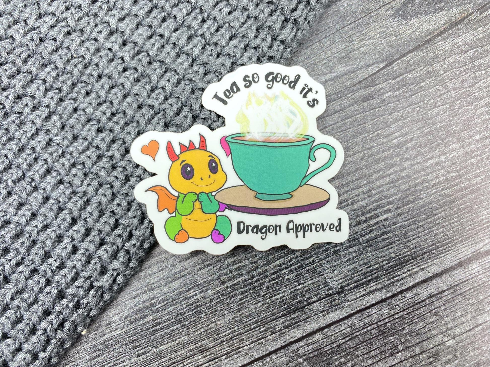 Die-cut vinyl sticker showing a yellow chibi dragon next to a steaming teal teacup, with the text "Tea so good it's dragon approved," on a gray knit and wood background
