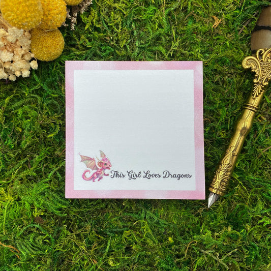 Square sticky note pad with a pink dragon illustration and the text 'This Girl Loves Dragons,' on a moss background with a quill pen