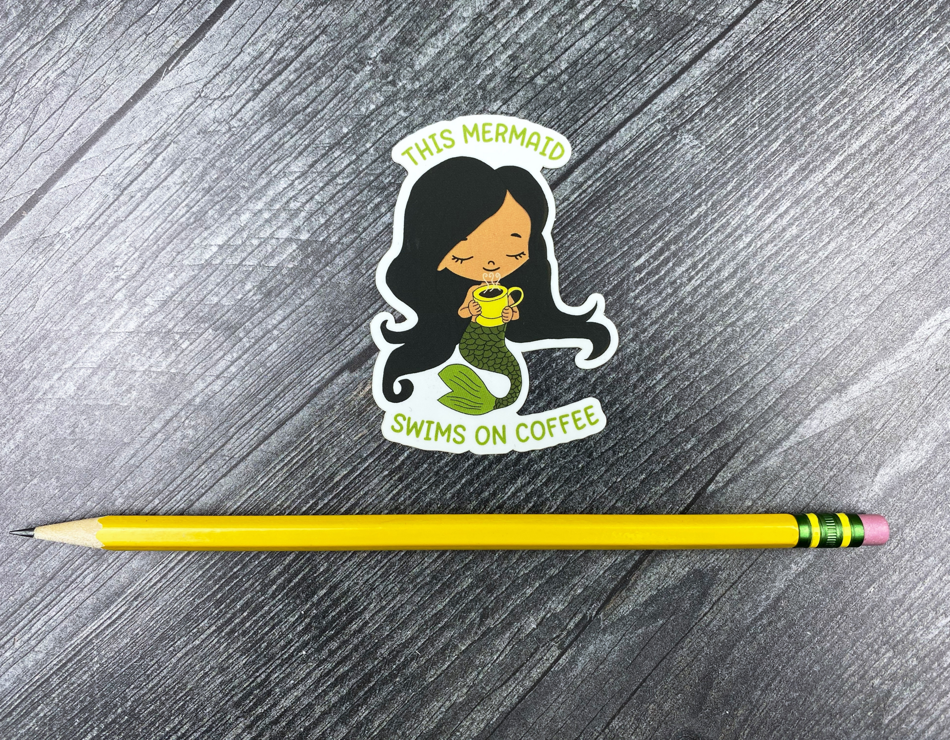 Die-cut vinyl sticker showing a dark-haired mermaid with a green tail holding a steaming yellow mug, with the text "This mermaid swims on coffee," on a gray wood background next to a yellow pencil