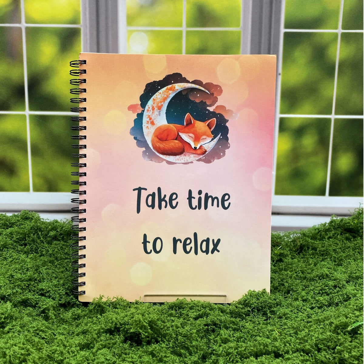 Spiral-bound notebook with a peach-to-pink gradient cover featuring a sleeping fox curled on a crescent moon, reading 'Take time to relax,' displayed on a moss mat with a window backdrop.