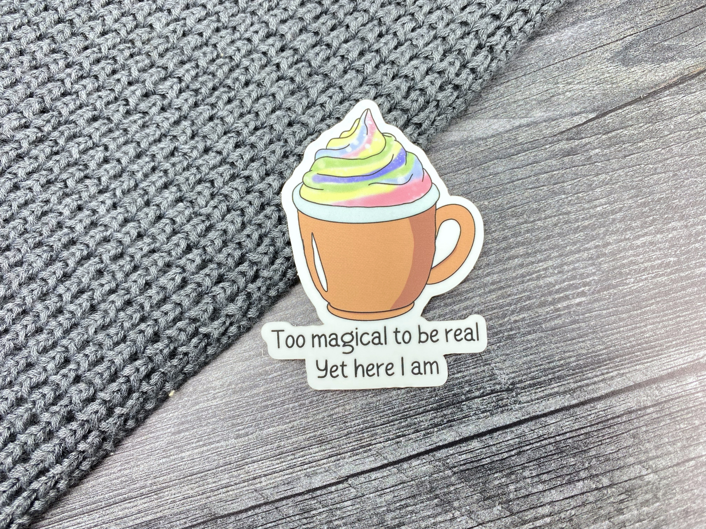 Die-cut vinyl sticker showing an orange mug topped with rainbow swirled whipped cream, with the text "Too magical to be real, yet here I am," on a gray knit and wood background