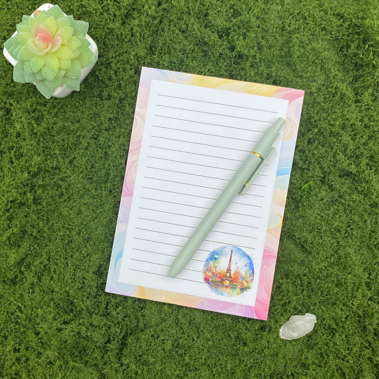 Lined notepad with a pastel swirl border and a circular watercolor illustration of the Paris skyline and Eiffel Tower, resting on artificial grass next to a sage green pen, small succulent, and crystal.