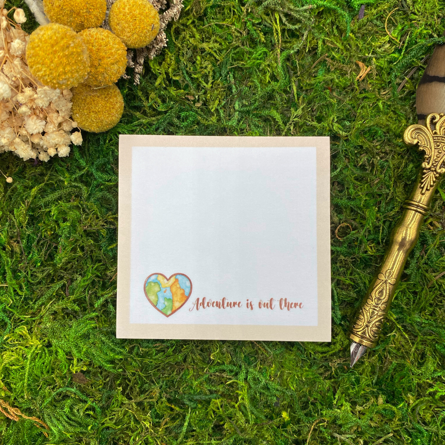 Square sticky note pad with a tan border, a heart-shaped watercolor world map, and the words "Adventure is out there," resting on moss next to dried yellow and white flowers and a gold quill pen.