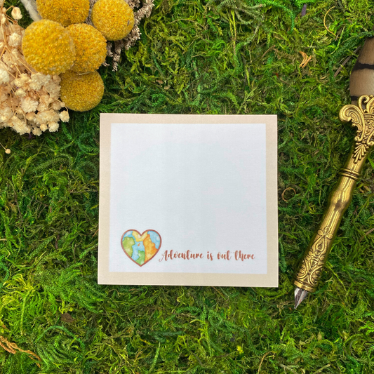 Square sticky note pad with a tan border, a heart-shaped watercolor world map, and the words "Adventure is out there," resting on moss next to dried yellow and white flowers and a gold quill pen.
