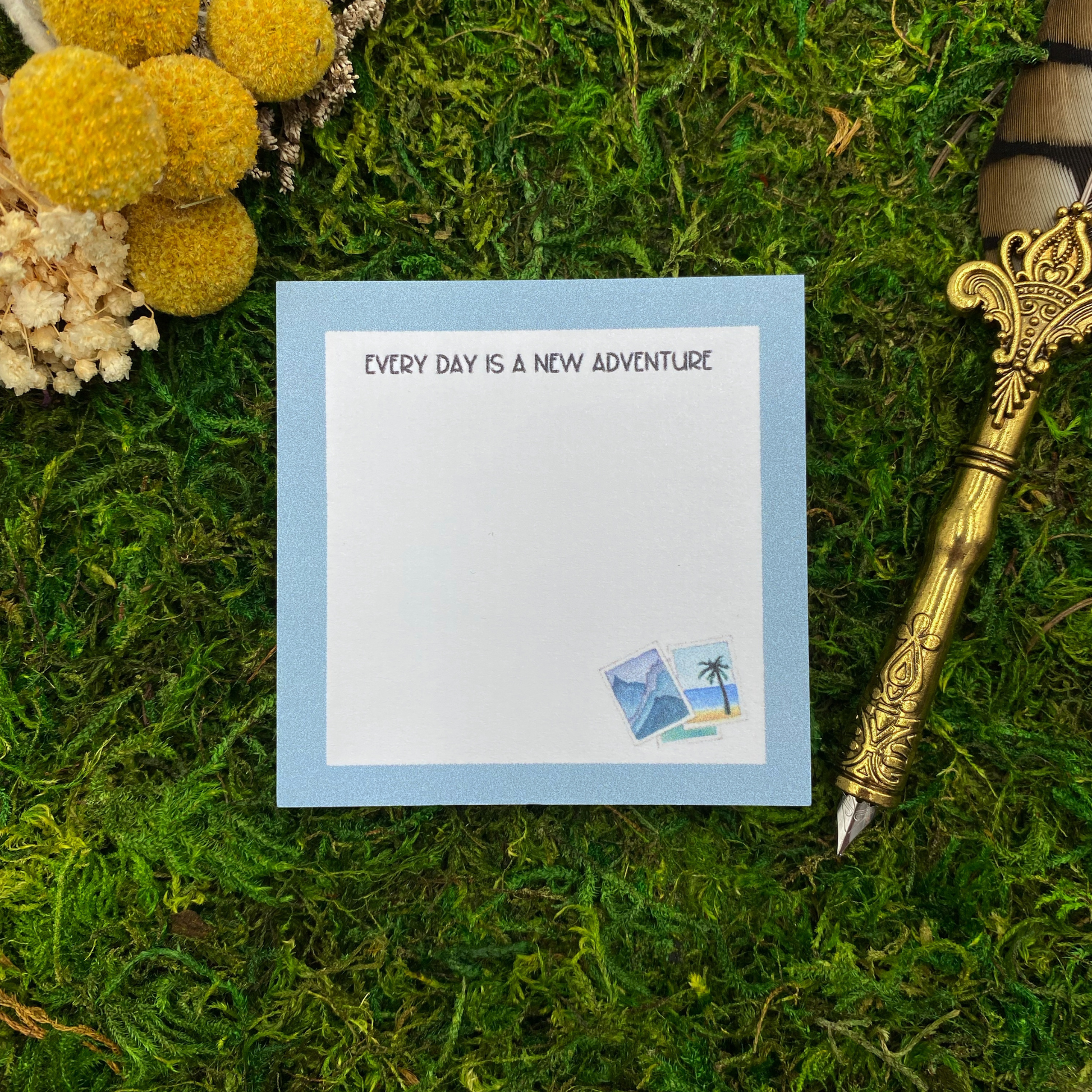 Square sticky note pad with a light blue border, small illustrated travel photos of mountains and a palm tree beach, and the words "Every day is a new adventure," resting on moss next to dried yellow and white flowers and a gold quill pen.