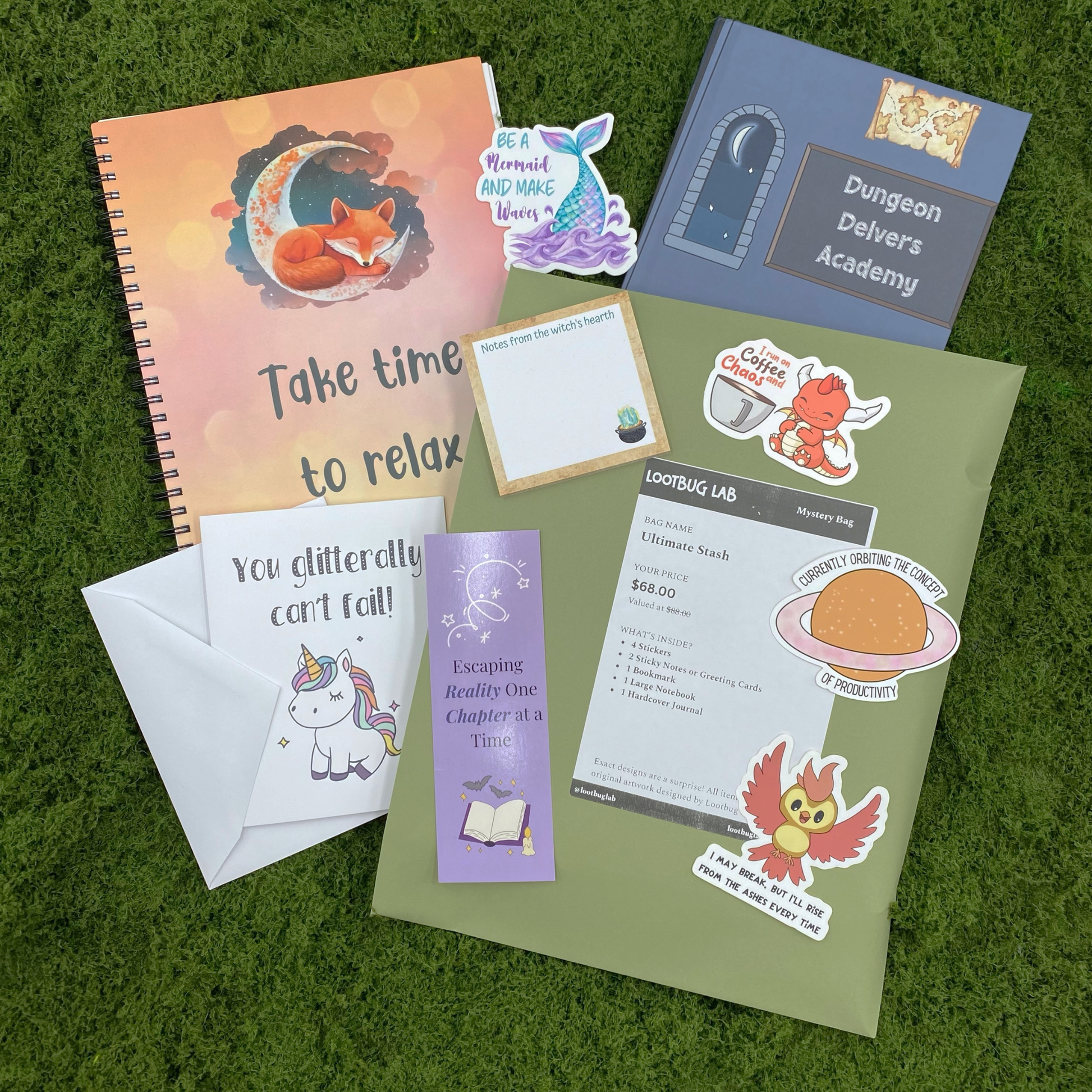 A Lootbug Lab ultimate stash mystery bag with a reveal card surrounded by sample items including stickers, a bookmark, a large notebook, a hardcover journal, and sticky note pads on a green moss surface