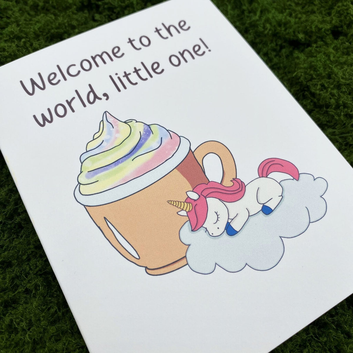 Close-up of the card's cover showing "Welcome to the world, little one!" above a sleeping pink-maned baby unicorn on a cloud next to a mug of rainbow whipped cream cocoa.
