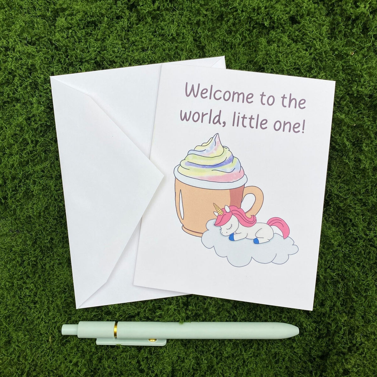 Folded greeting card reading "Welcome to the world, little one!" with a sleeping baby unicorn and cocoa mug illustration, resting on a white envelope on a mossy surface, with a sage green pen below.