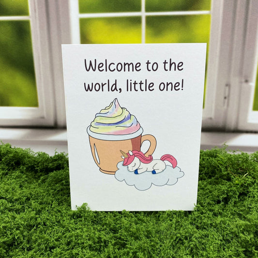 Greeting card that reads "Welcome to the world, little one!" above an illustration of a sleeping baby unicorn with pink mane curled on a cloud beside a mug of rainbow-swirled hot cocoa, standing upright on a moss surface in front of a white-framed window.