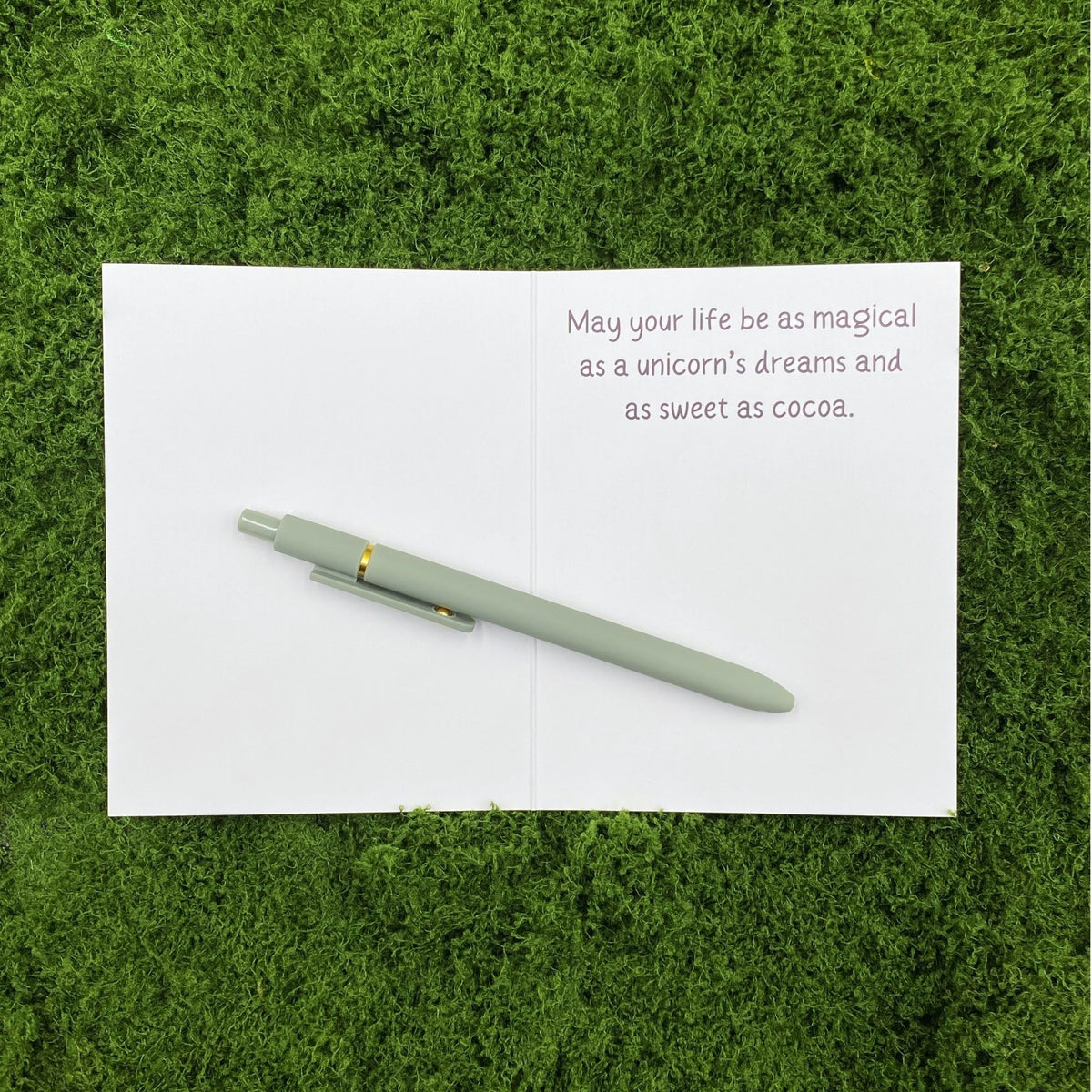 Card opened to show the printed message "May your life be as magical as a unicorn's dreams and as sweet as cocoa" on the right page, with a sage green pen laid across the blank left page on a mossy background.
