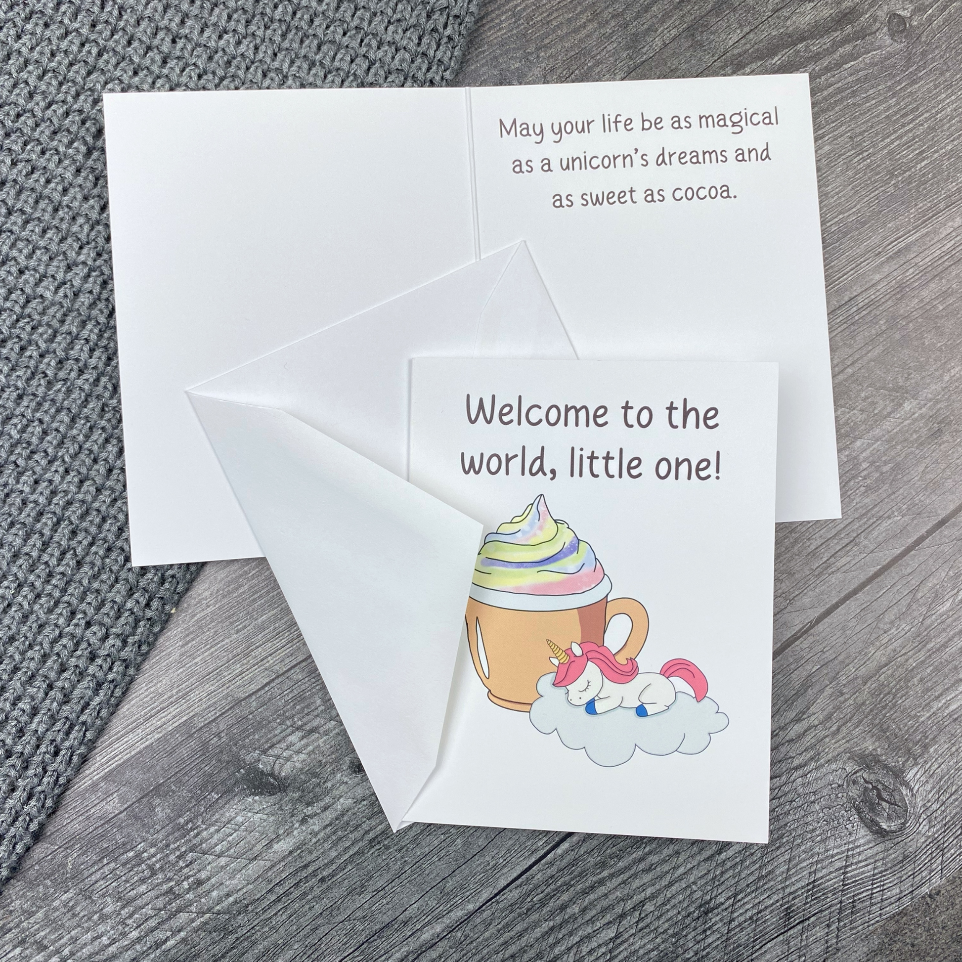 Greeting card lying open on a grey knit blanket and wood surface, showing the front with "Welcome to the world, little one!" and an illustration of a sleeping baby unicorn on a cloud beside a swirl-topped cocoa mug, next to the inside message "May your life be as magical as a unicorn's dreams and as sweet as cocoa," with the envelope tucked between the front and inside of the card.