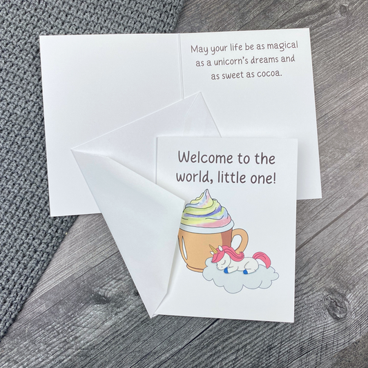 Greeting card lying open on a grey knit blanket and wood surface, showing the front with "Welcome to the world, little one!" and an illustration of a sleeping baby unicorn on a cloud beside a swirl-topped cocoa mug, next to the inside message "May your life be as magical as a unicorn's dreams and as sweet as cocoa," with the envelope tucked between the front and inside of the card.