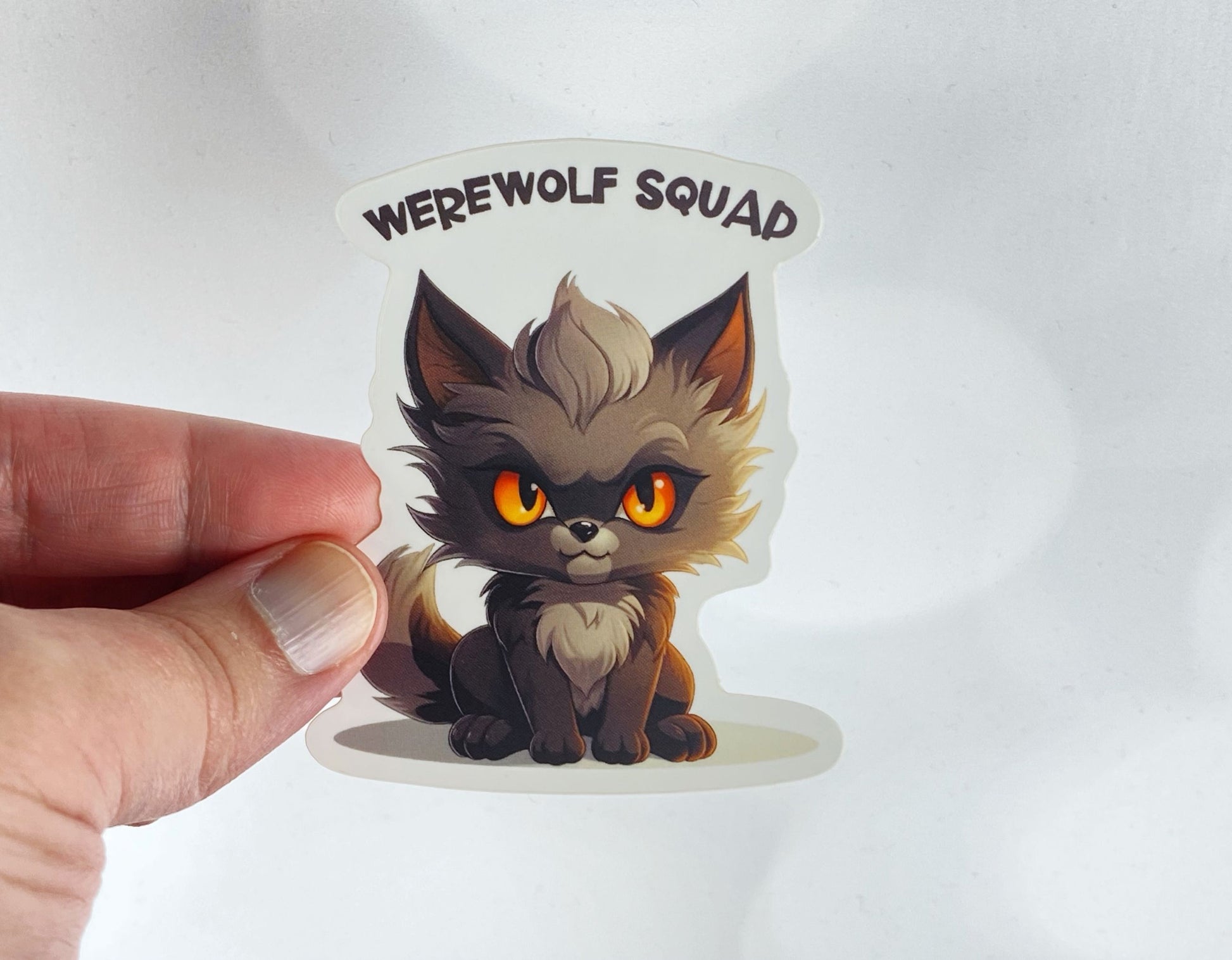 A hand holds a sticker illustration of a fierce, orange-eyed baby werewolf, reading "Werewolf Squad," against a white background.