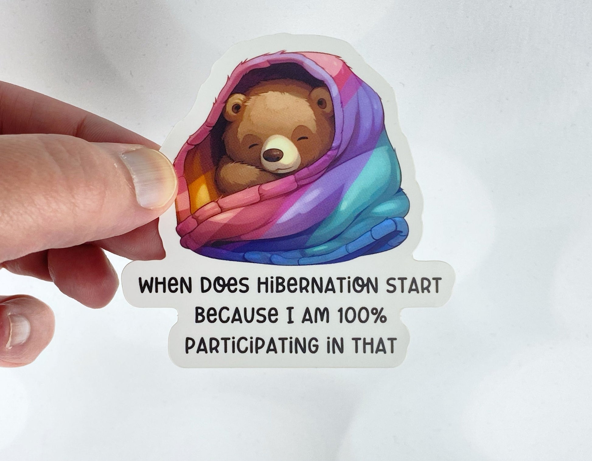 A hand holds a sticker illustration of a sleepy bear wrapped in a rainbow-striped blanket, reading "When does hibernation start because I am 100% participating in that," against a white background.