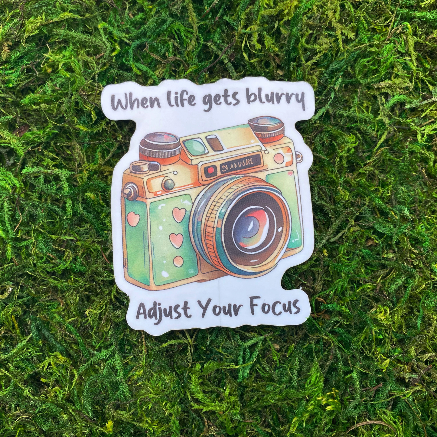A sticker illustration of a vintage camera with heart details, reading "When life gets blurry, adjust your focus," resting on artificial moss.