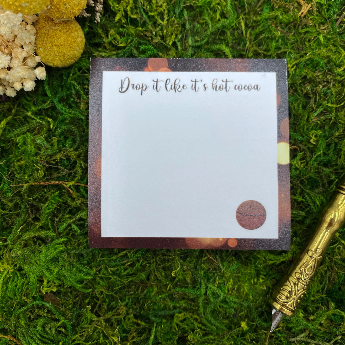 Square sticky note pad with a warm bokeh-light border, a small illustrated cocoa bomb, and the words "Drop it like it's hot cocoa," resting on moss next to dried yellow and white flowers and a gold quill pen.