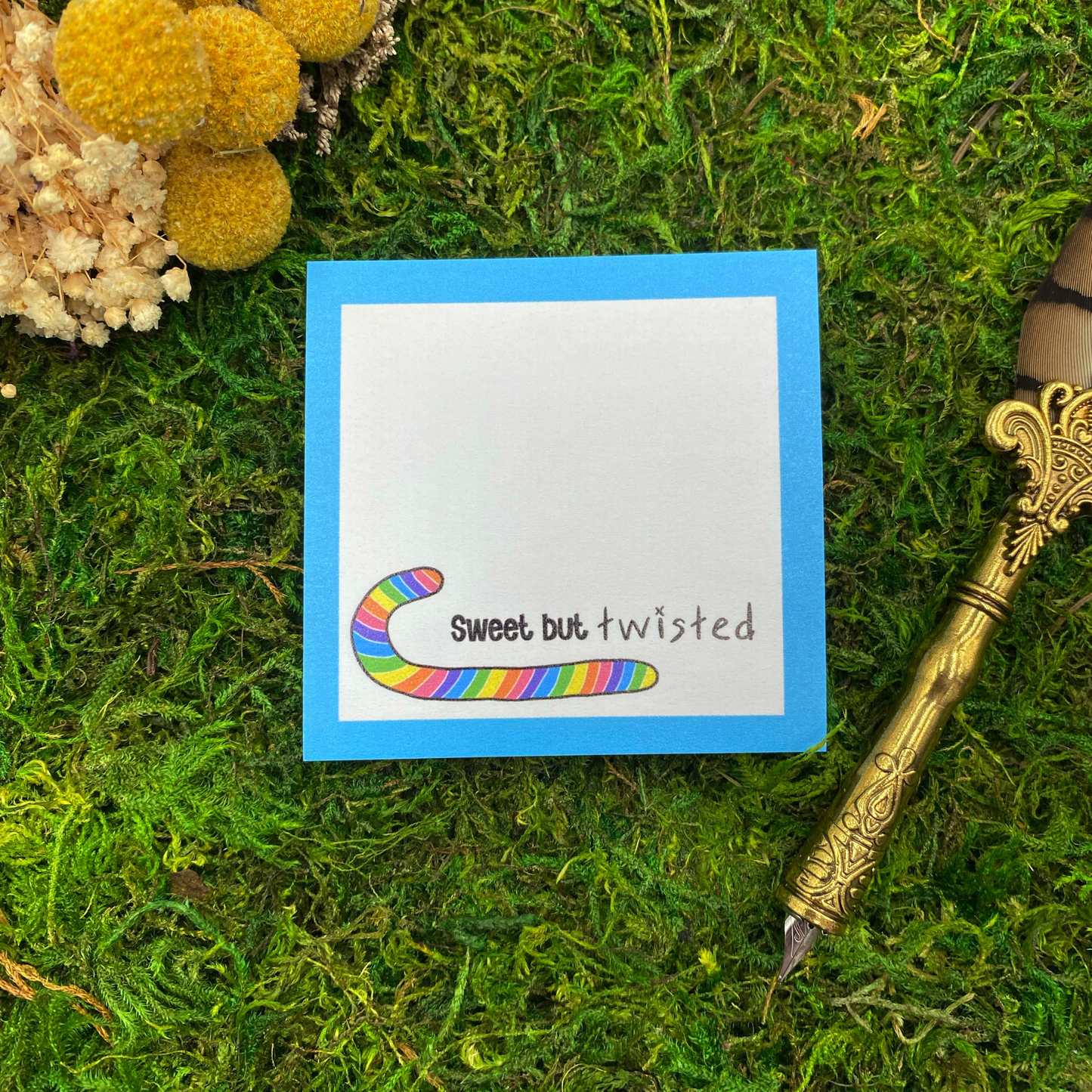 Square sticky note pad with a bright blue border, a rainbow-striped candy cane illustration, and the words "Sweet but twisted," resting on moss next to dried yellow and white flowers and a gold quill pen.