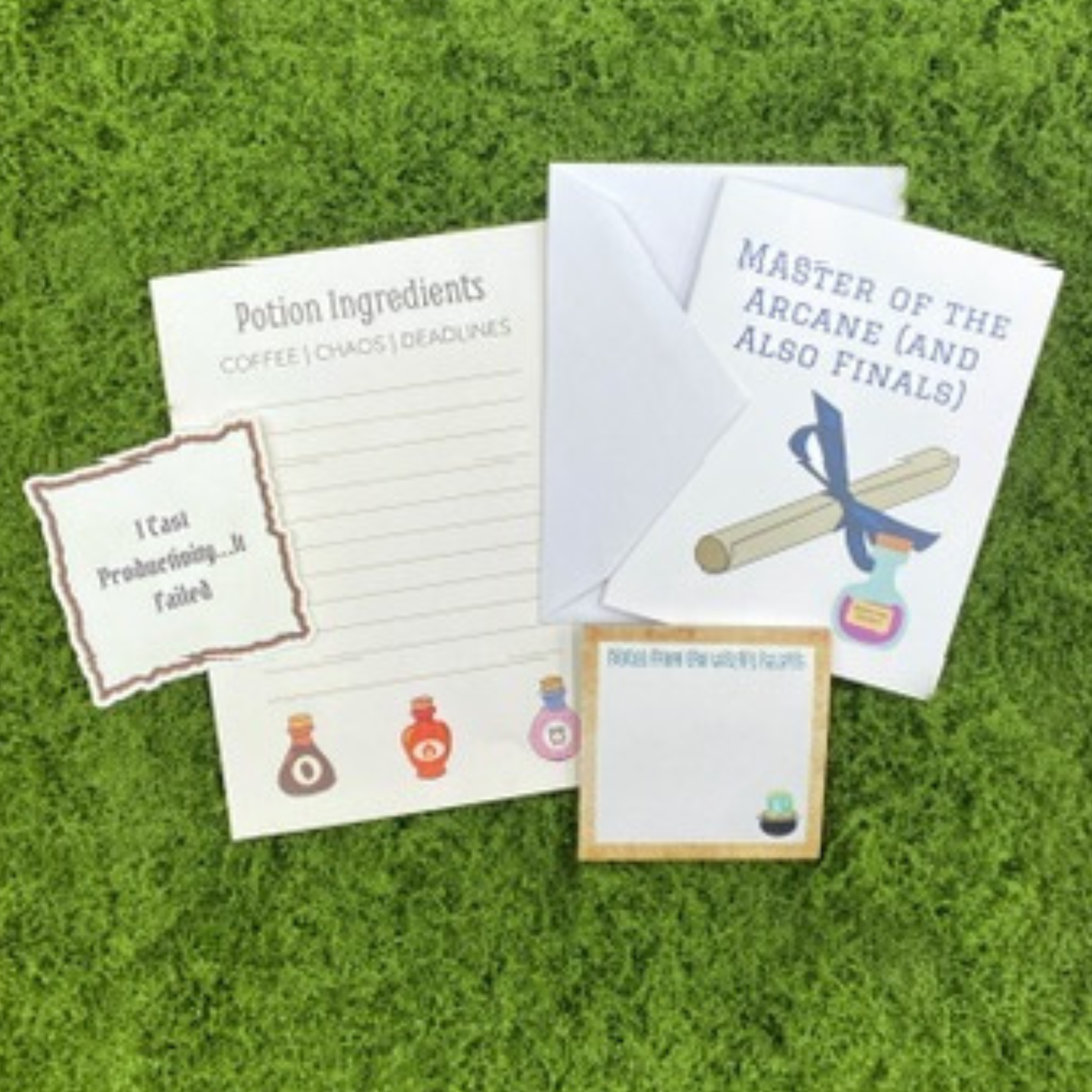 A curated gift set on artificial grass featuring a "Potion Ingredients" notepad, a sticky note reading "I Cast Productivity...It Failed," a witch's cauldron sticky note, and a greeting card that reads "Master of the Arcane (and Also Finals)" with a diploma illustration.