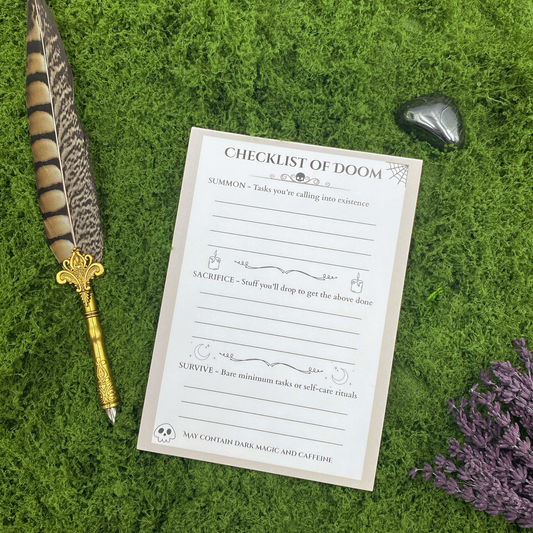 Notepad headed "Checklist of Doom" with three sections labeled Summon, Sacrifice, and Survive, decorated with a spiderweb, skull, candles, and crescent moons, resting on artificial grass next to a gold quill pen, an obsidian stone, and dried lavender.