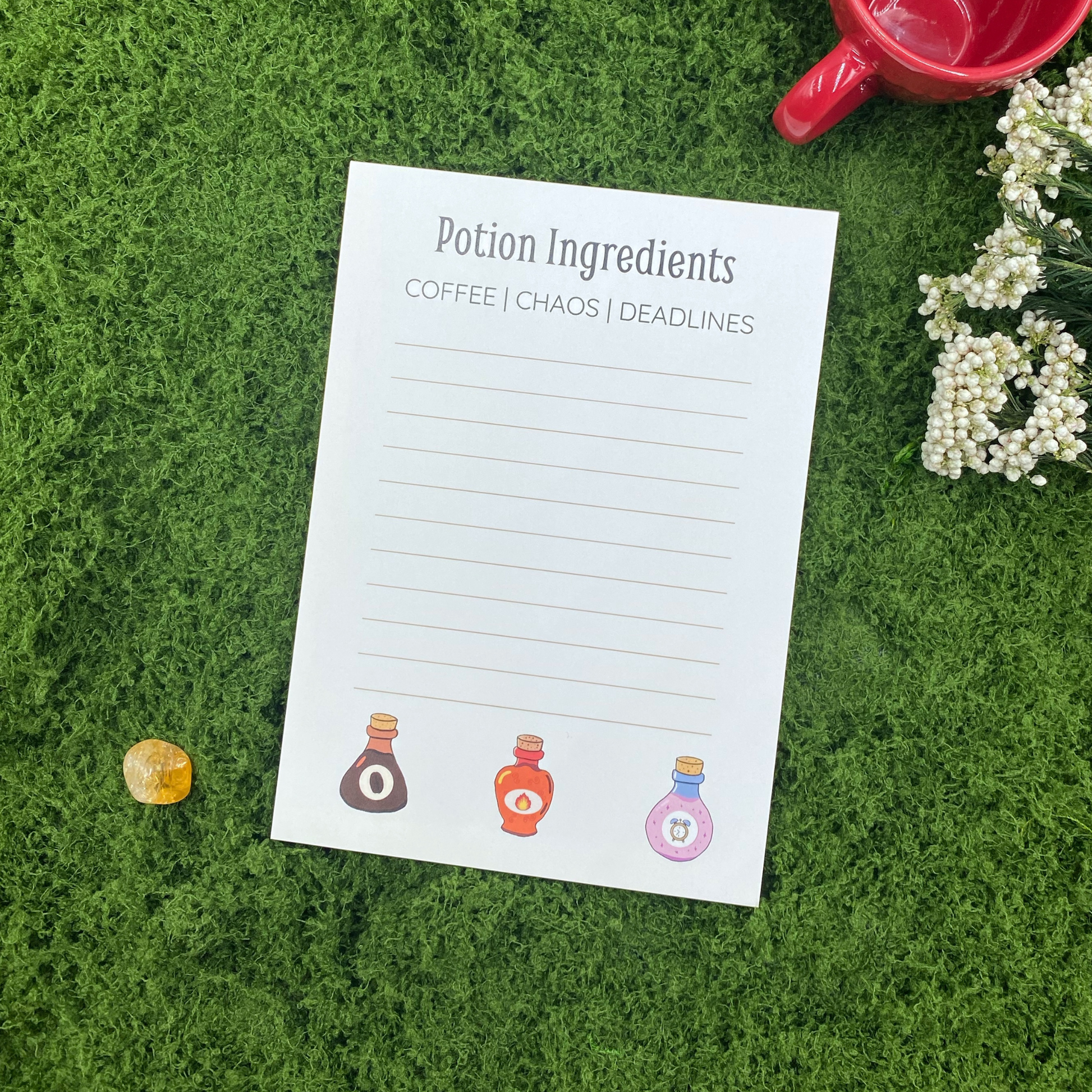 Notepad headed "Potion Ingredients: Coffee, Chaos, Deadlines" with three illustrated potion bottles along the bottom, resting on artificial grass next to a red mug, a citrine crystal, and white baby's breath flowers.
