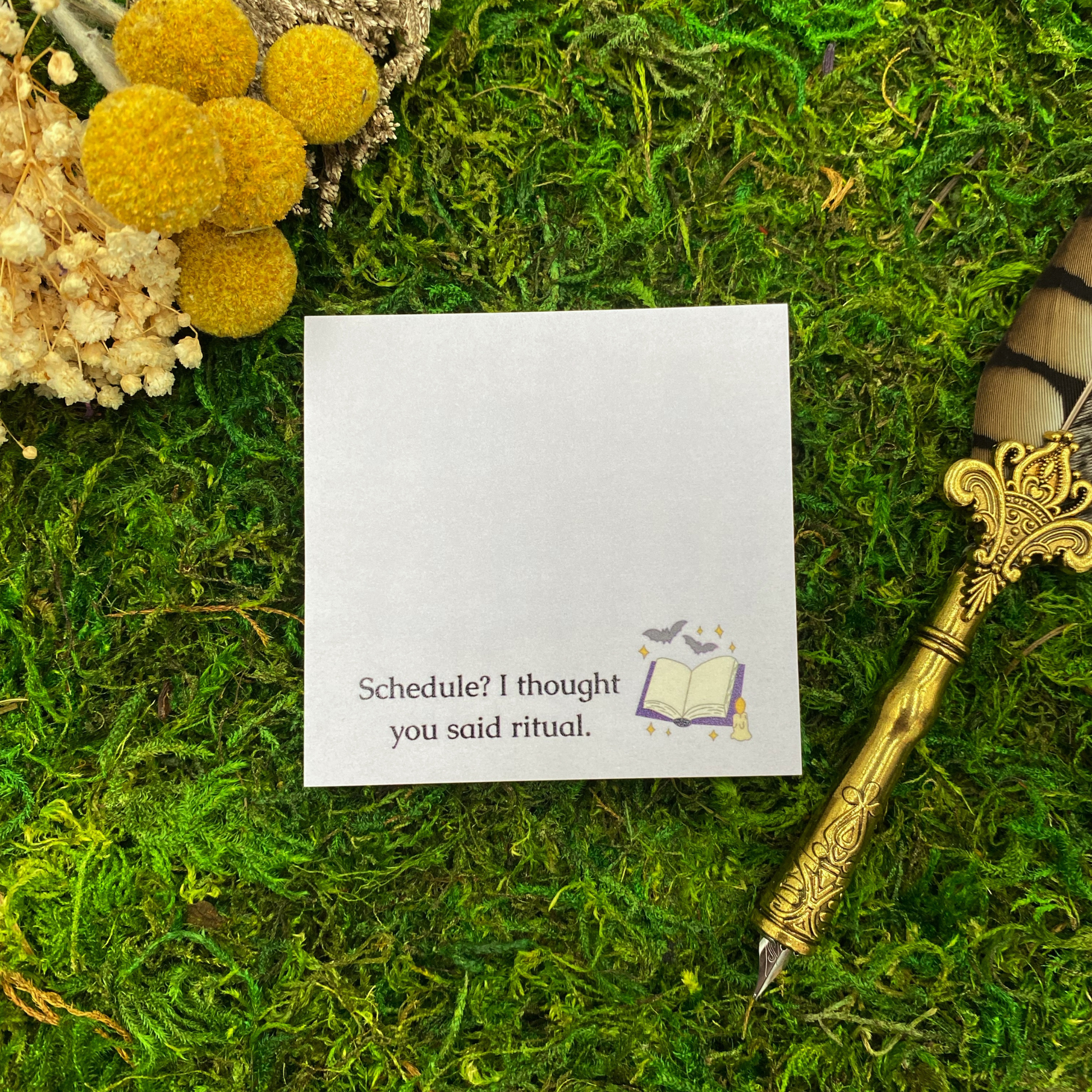 Square sticky note pad with a small illustrated open spellbook, candle, and flying bats, and the words "Schedule? I thought you said ritual," resting on moss next to dried yellow and white flowers and a gold quill pen.