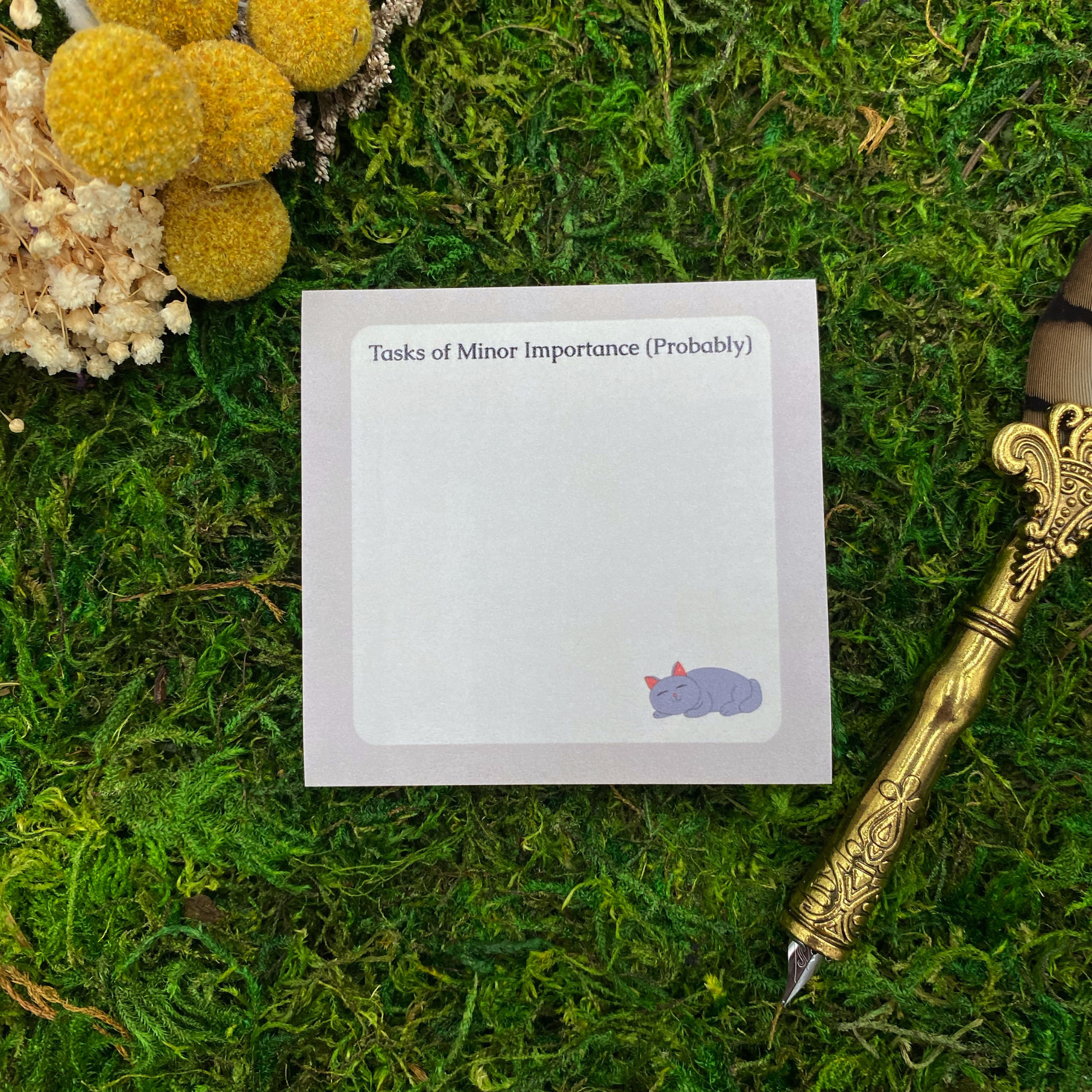 Square sticky note pad with a lavender border, a sleeping grey cat illustration, and the words "Tasks of Minor Importance (Probably)," resting on moss next to dried yellow and white flowers and a gold quill pen.