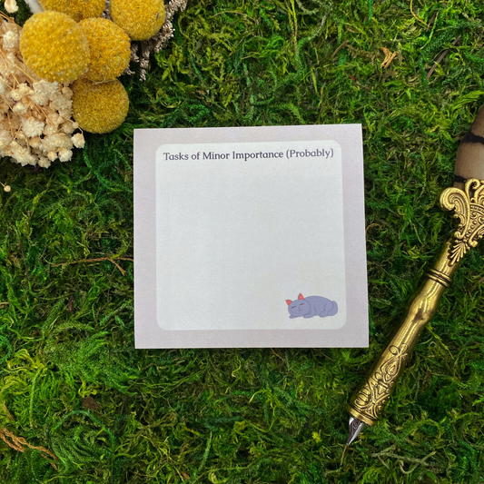 Square sticky note pad with a lavender border, a sleeping grey cat illustration, and the words "Tasks of Minor Importance (Probably)," resting on moss next to dried yellow and white flowers and a gold quill pen.